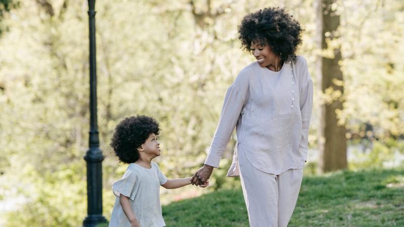 A mother and her young son walk hand in hand along a tree-lined path, sharing a smile.